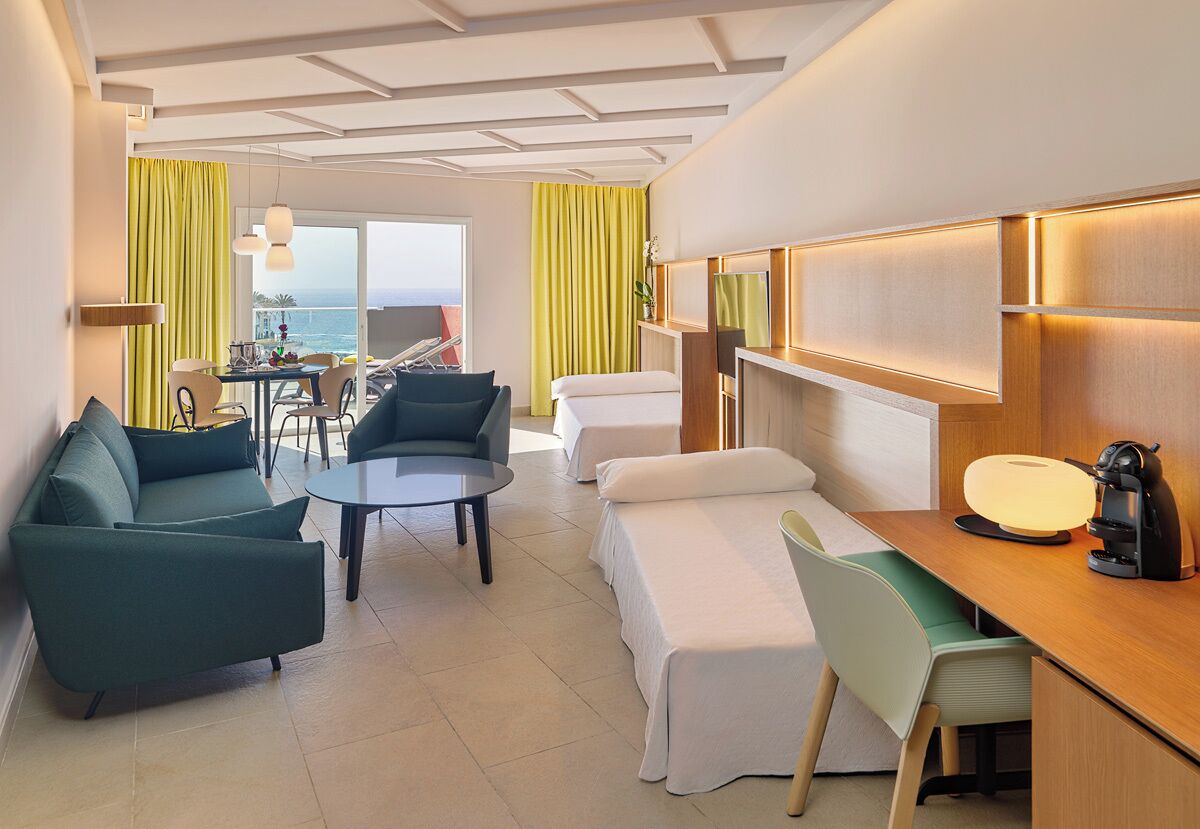 Habitación de hotel moderna con sofá, una mesa de comedor, escritorio con lámpara, cama y vistas al mar.