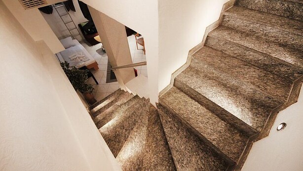 Un escalier dont les marches sont recouvertes d’une pierre à texture particulière, menant à une pièce où se trouvent un lit et une plante en pot.