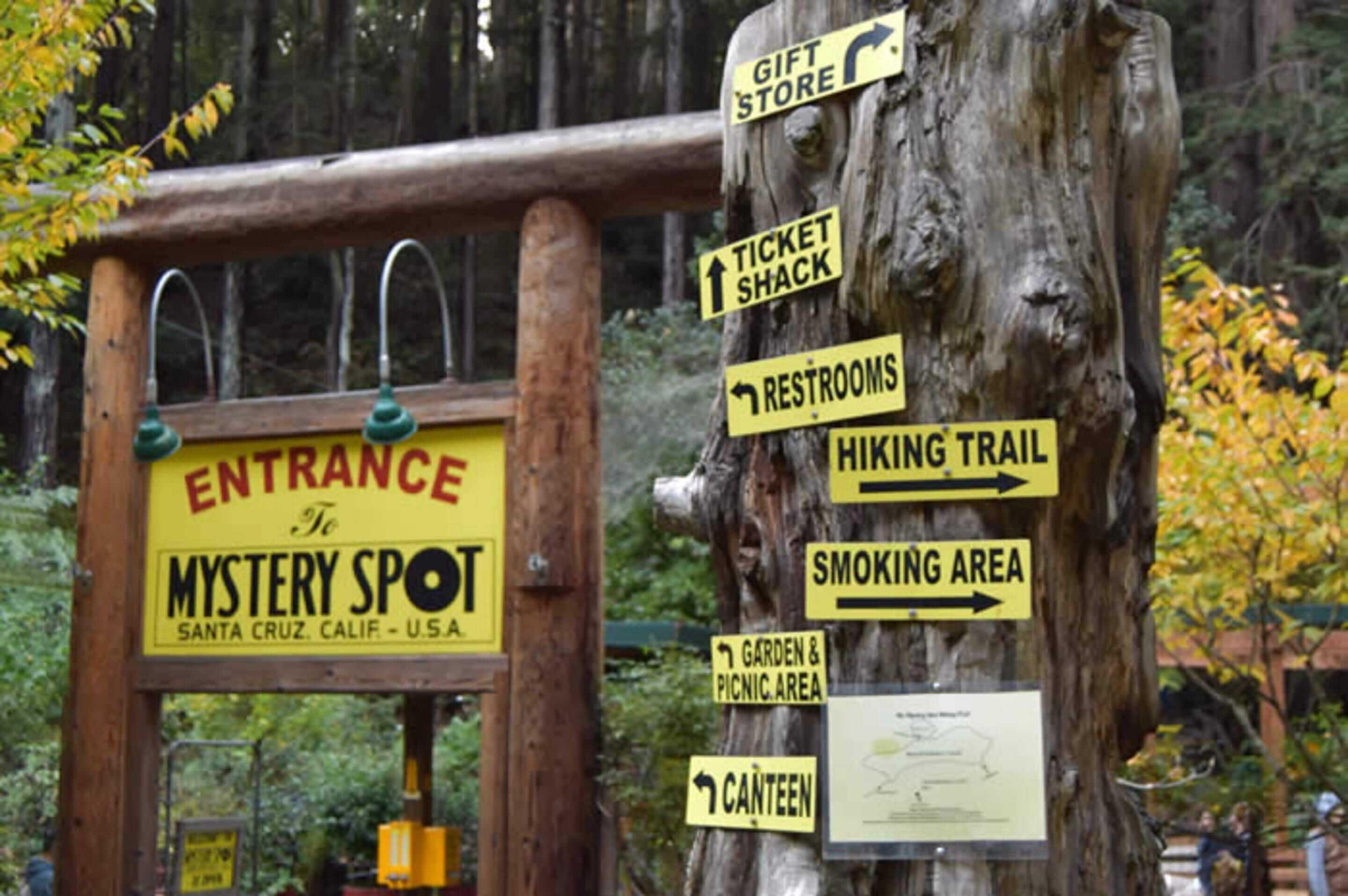Un cartel de madera con múltiples indicaciones direccionales para un parque, incluyendo la entrada a Mystery Spot, una tienda de regalos, una taquilla, baños, un sendero para caminatas, una zona para fumadores, un jardín y área de picnic, y un quiosco.