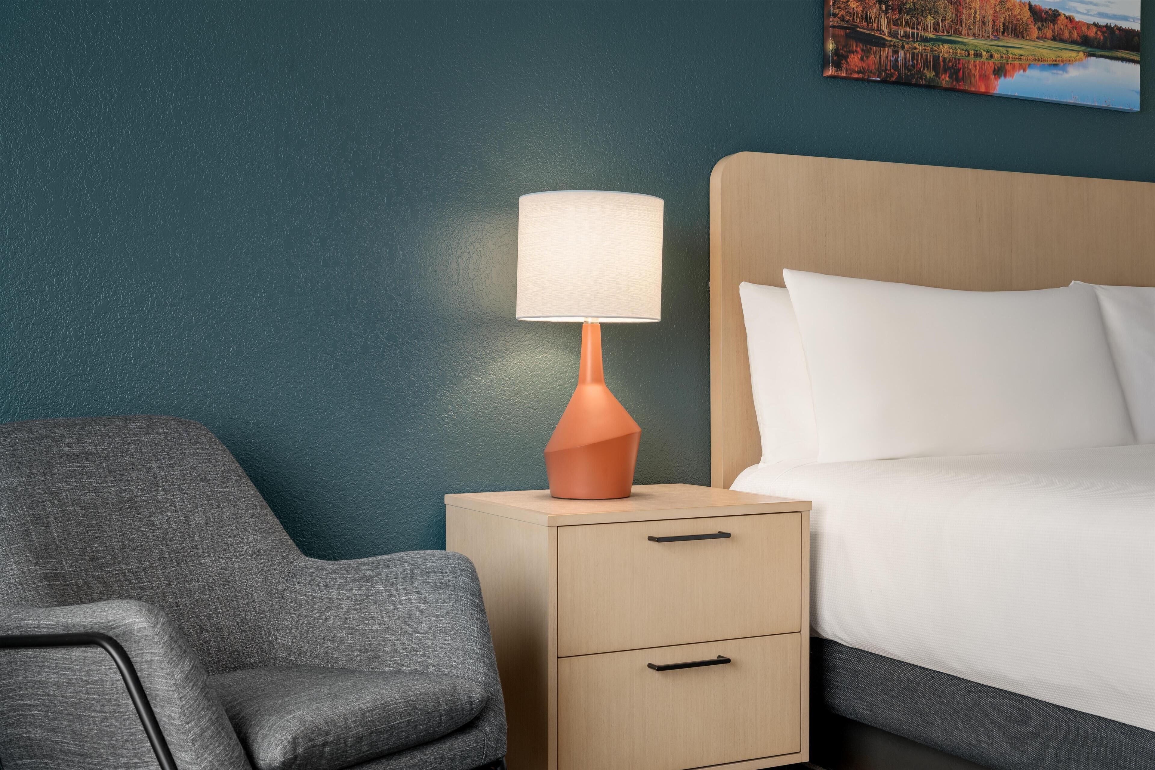 Una habitación de hotel con cama, mesita de noche con lámpara, una silla y un cuadro en la pared.