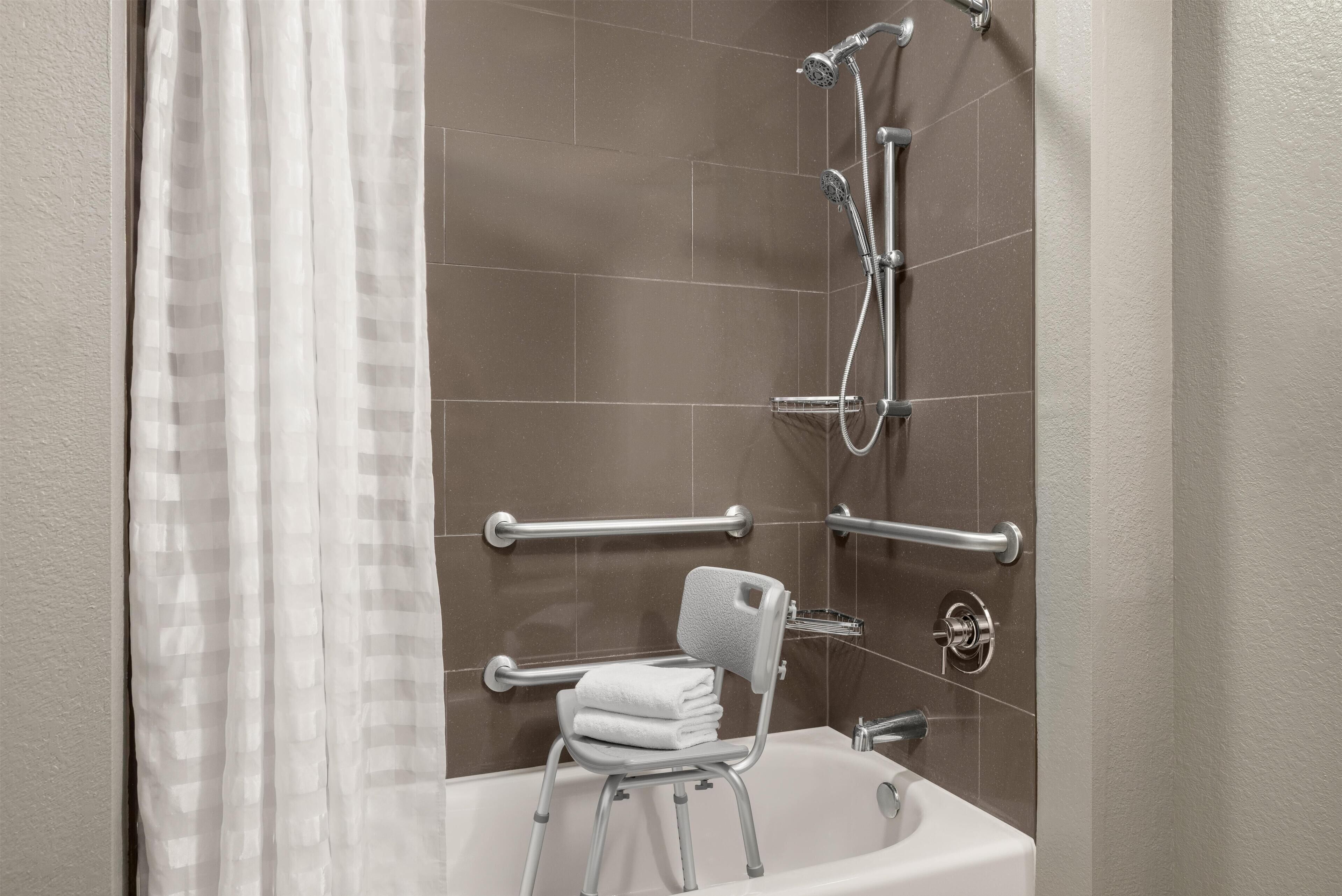 Un baño con bañera, ducha, barras de apoyo y una silla con toalla.