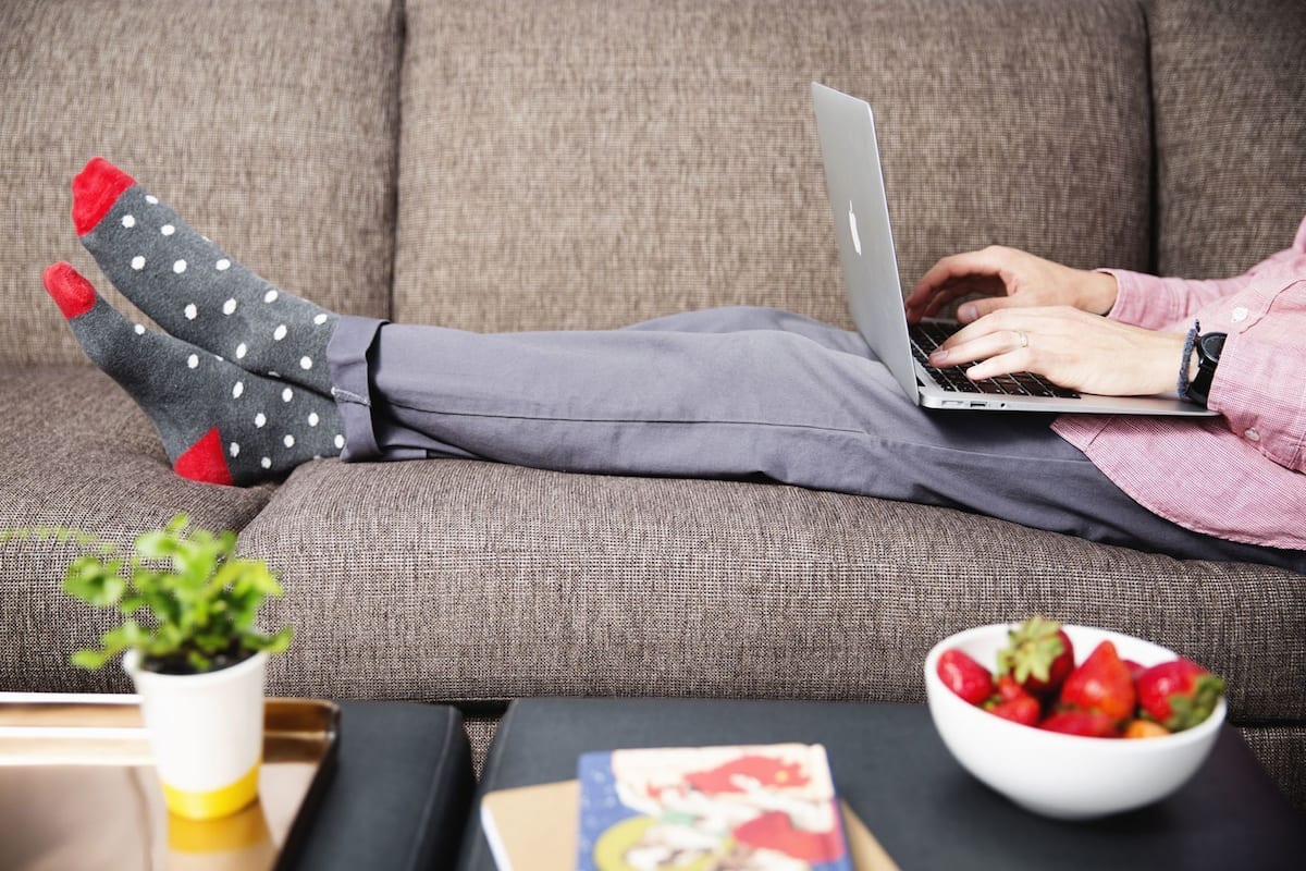 A person using a laptop on a couch with a bowl of strawberries and a book on a table.