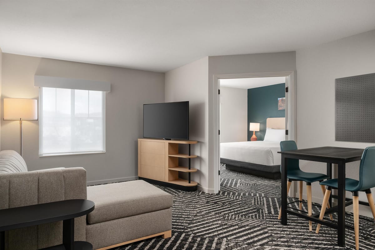 A hotel room with a sofa, a TV on a wooden stand, a dining table with chairs, a bed, and a patterned carpet.