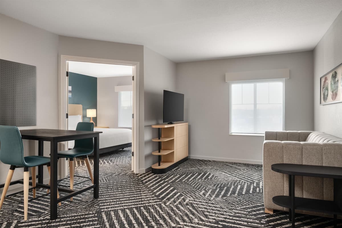 A hotel room with a bed, a desk with chairs, a TV on a stand, a sofa, a window, and a patterned carpet.