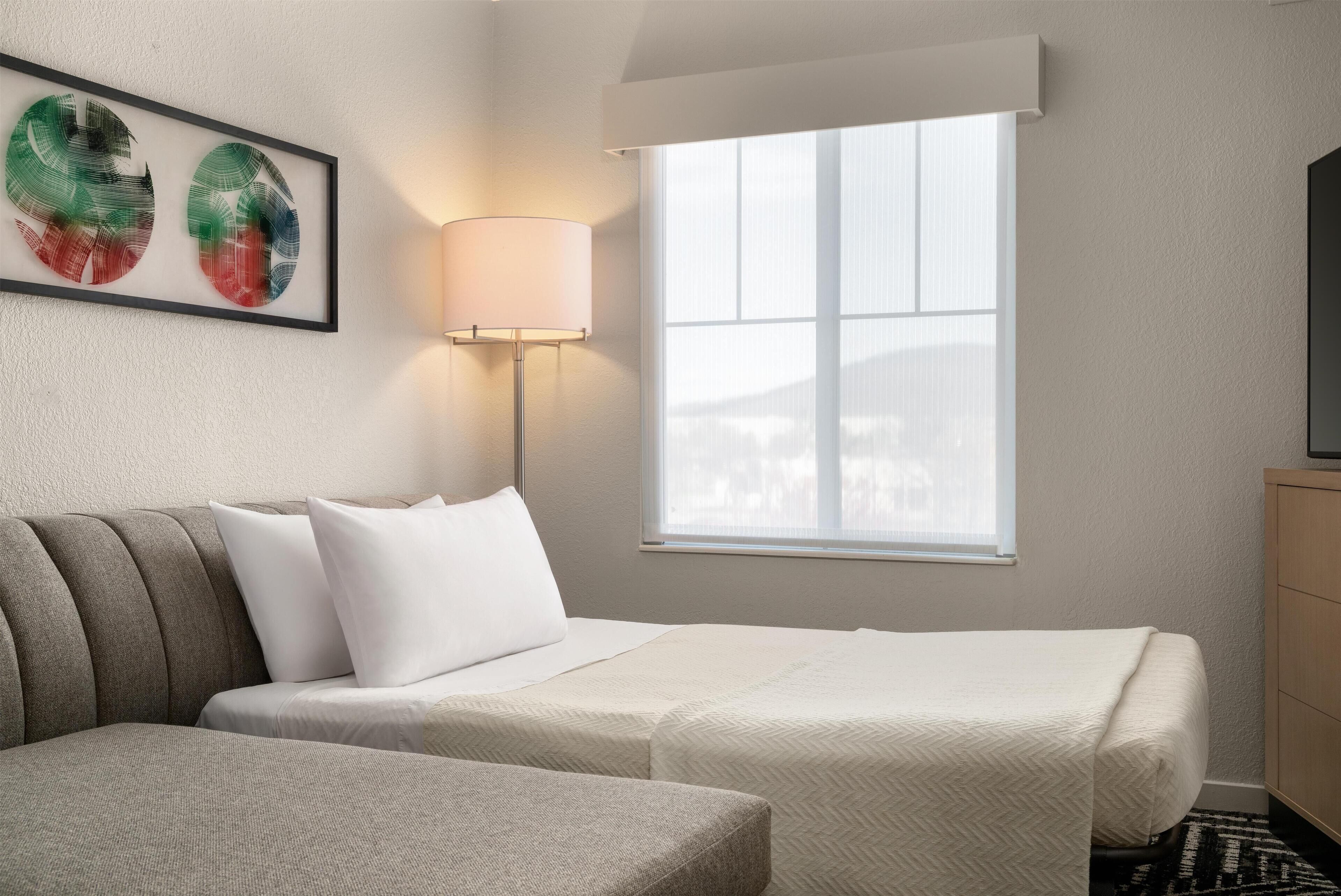 Ein Hotelzimmer mit zwei Betten, einem Fenster mit Jalousien, einer Lampe und einem gerahmten Kunstwerk an der Wand.
