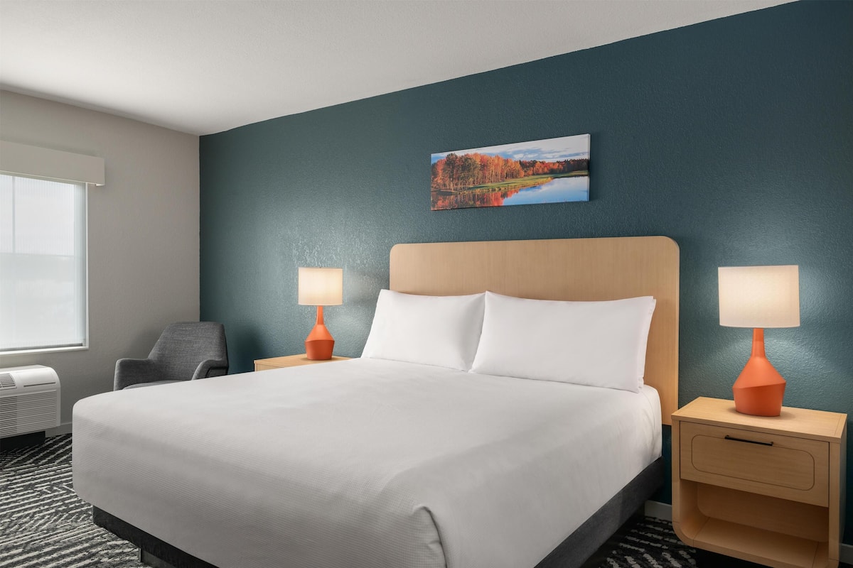 A hotel room with a large bed, two bedside lamps, a chair, a nightstand, a window, and a painting on the wall.