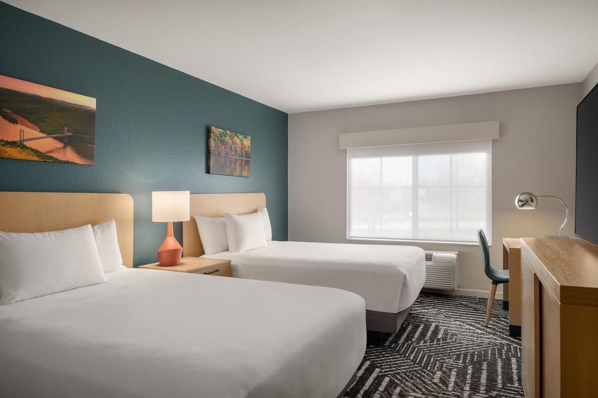 A hotel room with two beds, a desk, a chair, a window with blinds, and artwork on the walls.