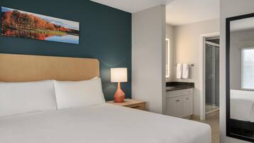 A hotel room with a bed, a nightstand with a lamp, a painting of a landscape, a bathroom with a shower, and a mirror.
