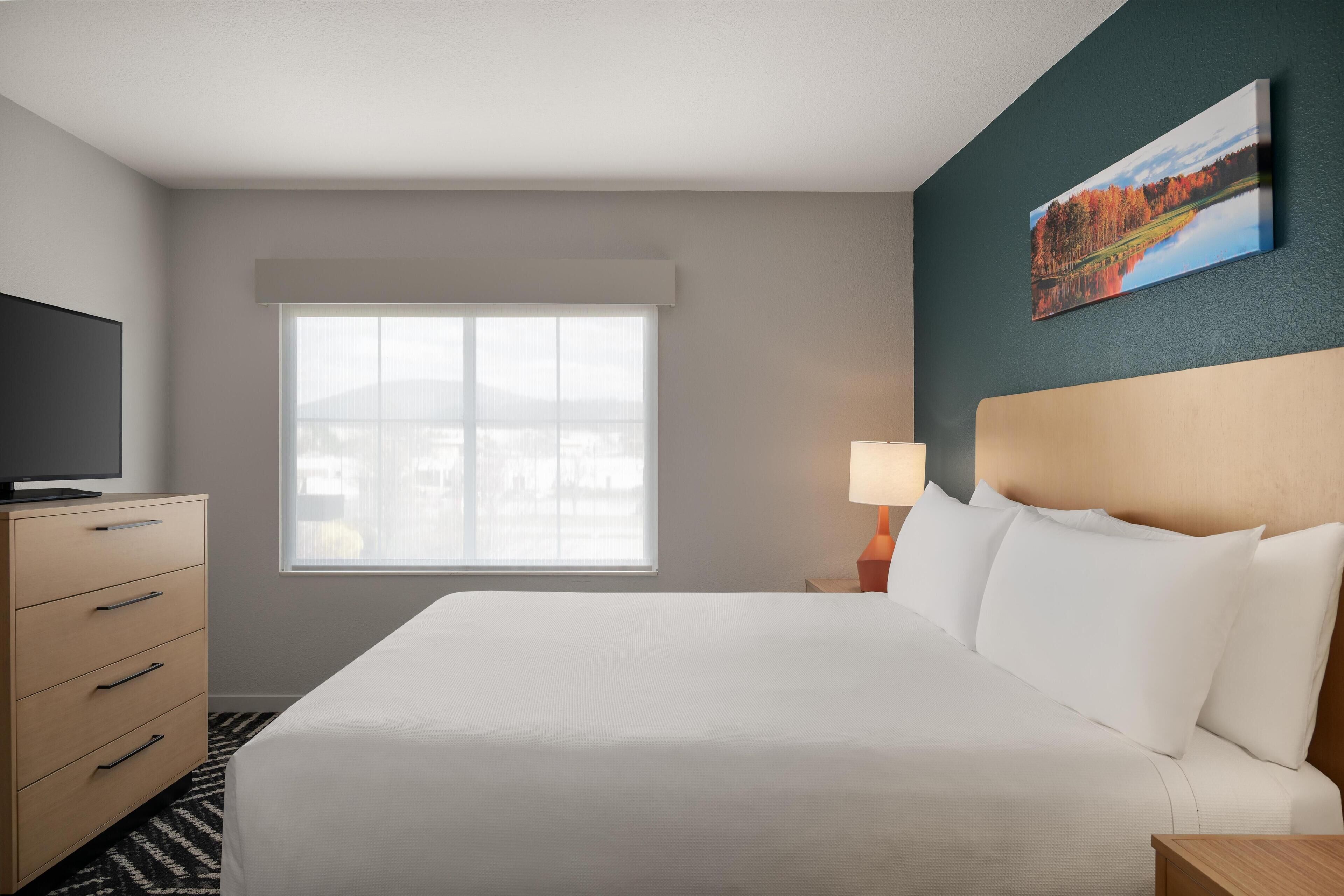 A hotel room with a bed, a TV, a dresser, a window, and a painting on the wall.