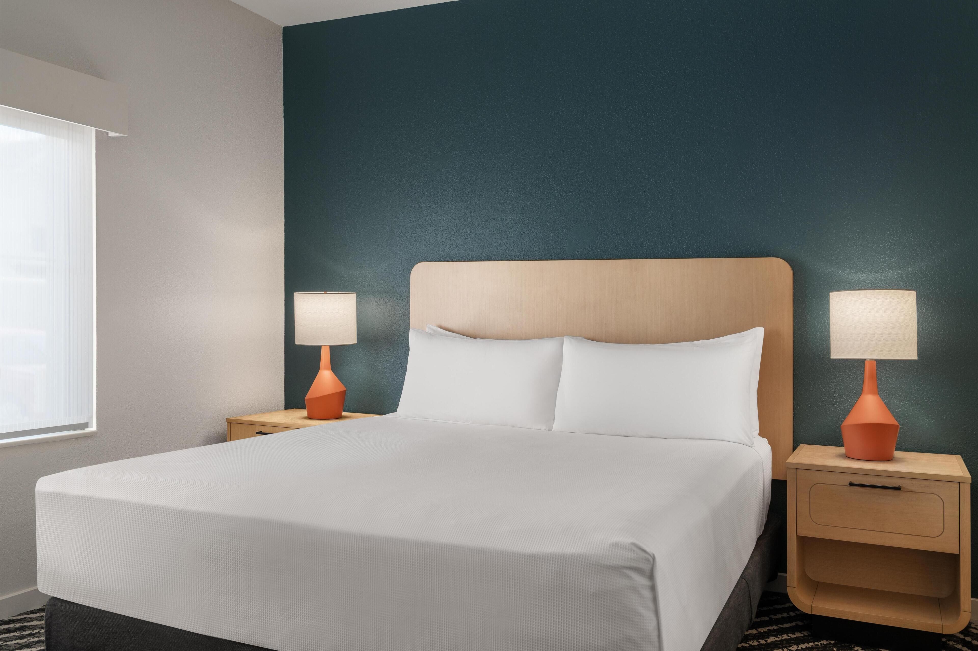 Una cama bien hecha con sábanas blancas, dos mesitas de noche con lámparas y una pared de color verde azulado oscuro.