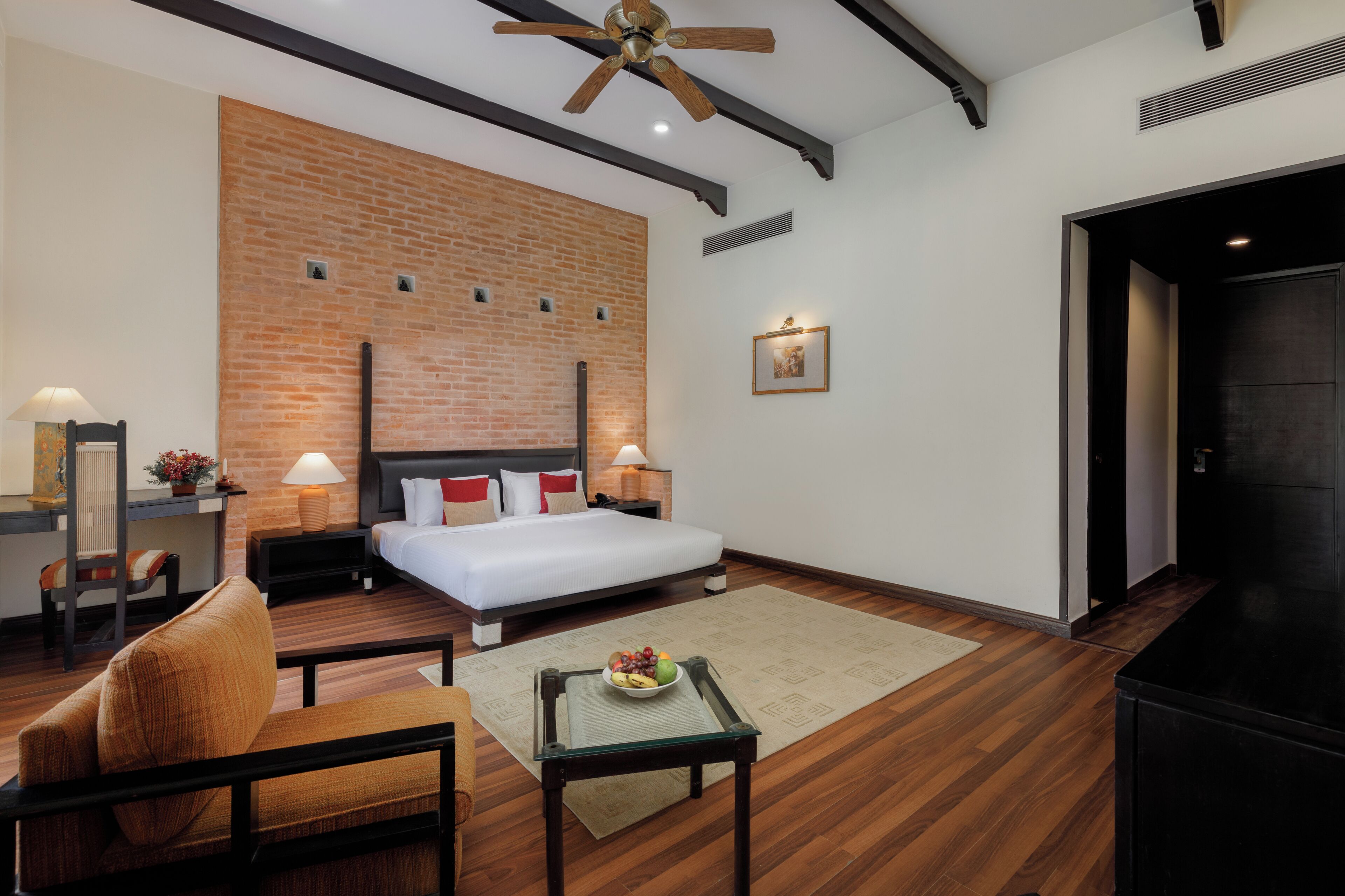 Una habitación de hotel moderna con una pared de acento de ladrillo, una cama grande, un escritorio con silla, una mesita con fruta y un ventilador de techo.