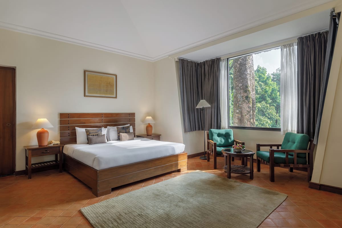 Ein Hotelzimmer mit einem großen Bett, zwei Nachttischen mit Lampen, einem grünen Sessel, einem kleinen Tisch ebenfalls mit einer Lampe und einem Fenster mit Vorhängen.