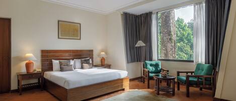 Habitación de hotel con una cama grande, dos mesitas de noche con lámparas, un sillón verde, una mesita pequeña con lámpara y una ventana con cortinas.