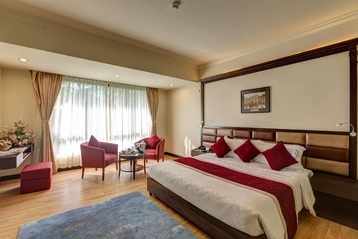 Ein Hotelzimmer mit einem großen Bett, zwei roten Stühlen, einem kleinen Tisch, einem Schreibtisch mit einer Lampe und einem fensterbepflanzten Fenster.