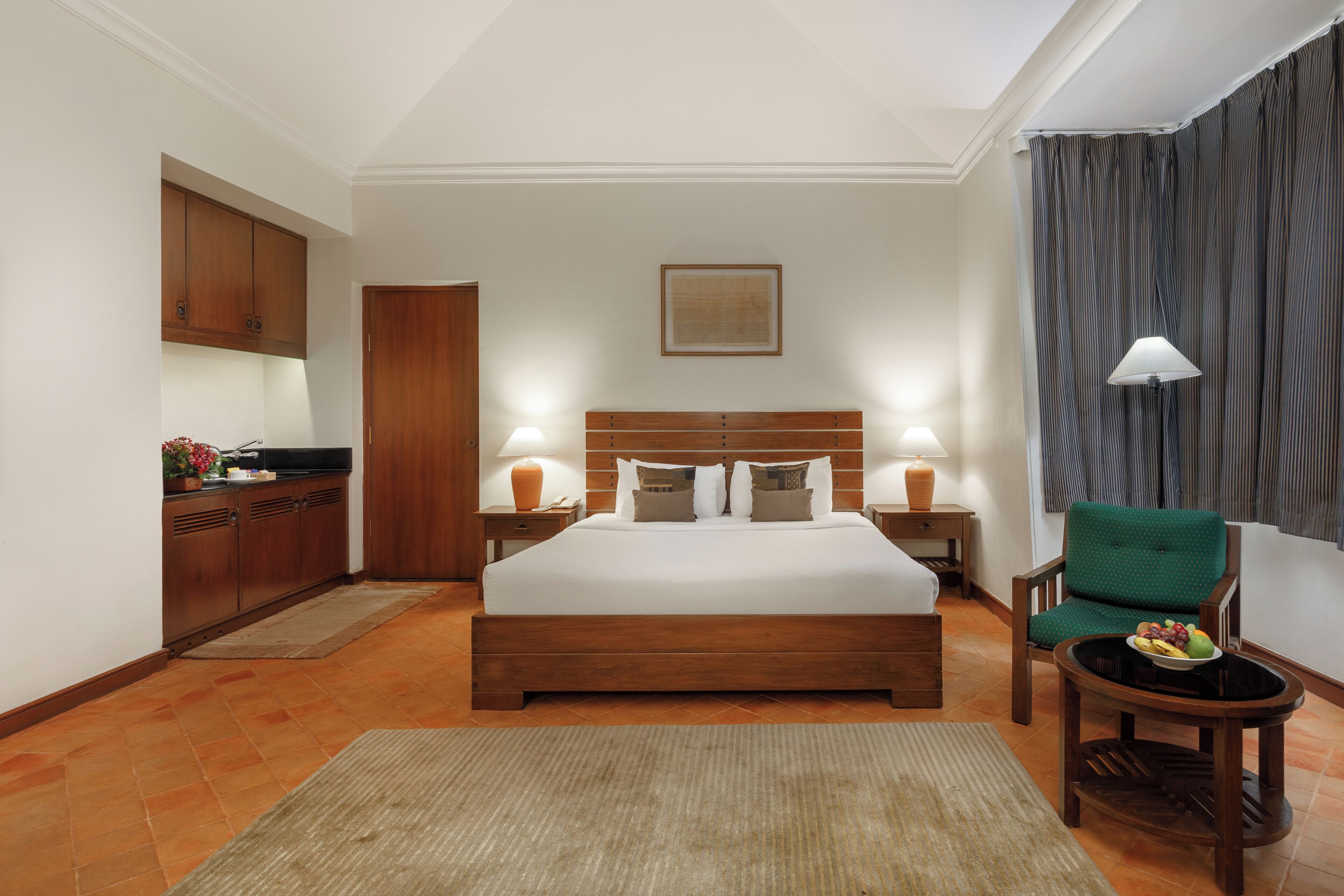 Een hotelkamer met een groot bed, twee nachtkastjes met lampen, een groene fauteuil, een klein tafeltje met fruit, en een kitchenette met kastjes en een spoelbak.