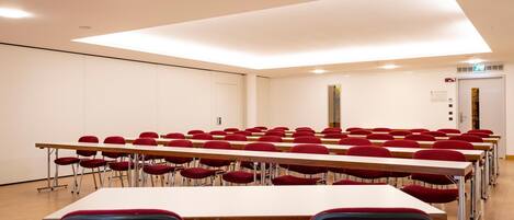 Un salón de conferencias con mesas y sillas dispuestas en filas, una lámpara en el techo y una puerta con letrero de salida de emergencia.