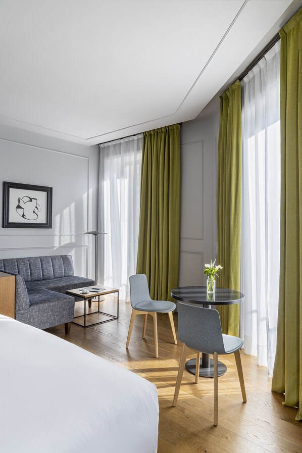 A modern hotel room with a bed, a gray sofa, a round table with two chairs, and a vase with flowers. - Tótem Madrid Hotel (Madrid)