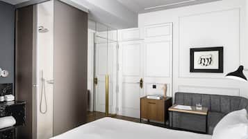 Una habitación de hotel moderna con una cama grande, una mesita de noche con lámpara, un pequeño tocador de madera, un cuadro enmarcado en la pared y un vestidor con ducha.