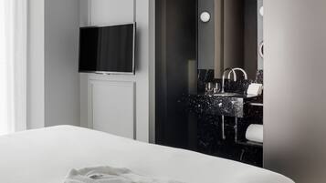 Una habitación de hotel moderna con una cama grande, un televisor de pantalla plana montado en la pared, un baño con encimera de mármol oscuro y un espejo amplio.