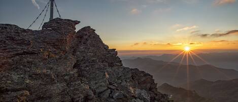 A rugged mountain landscape with a cross on a tripod at the peak, overlooking a valley with distant mountains and a sunset.