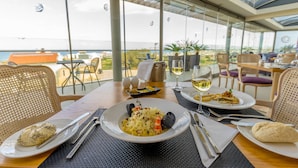 Uma mesa de jantar com pratos de comida, talheres e copos de vinho, em um restaurante com vista para o mar.