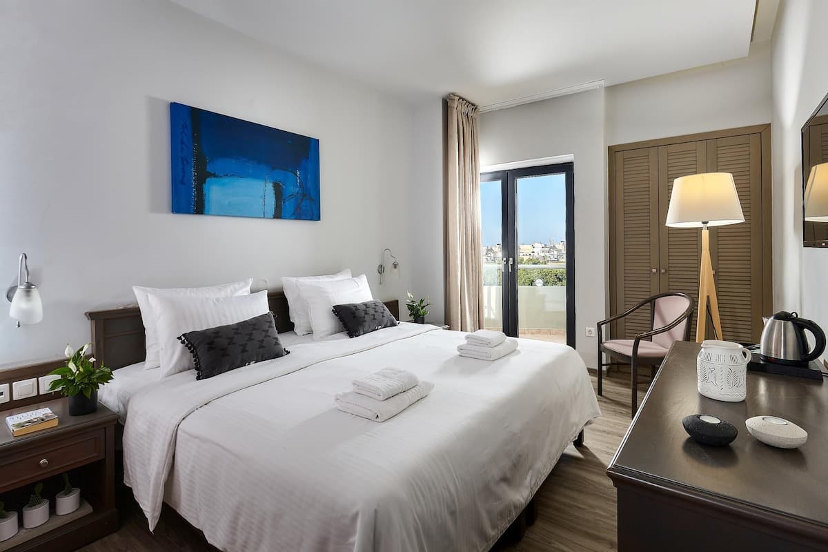 Una habitación de hotel con una cama grande, un escritorio con lámpara, cafetera y vistas a la ciudad desde la ventana.