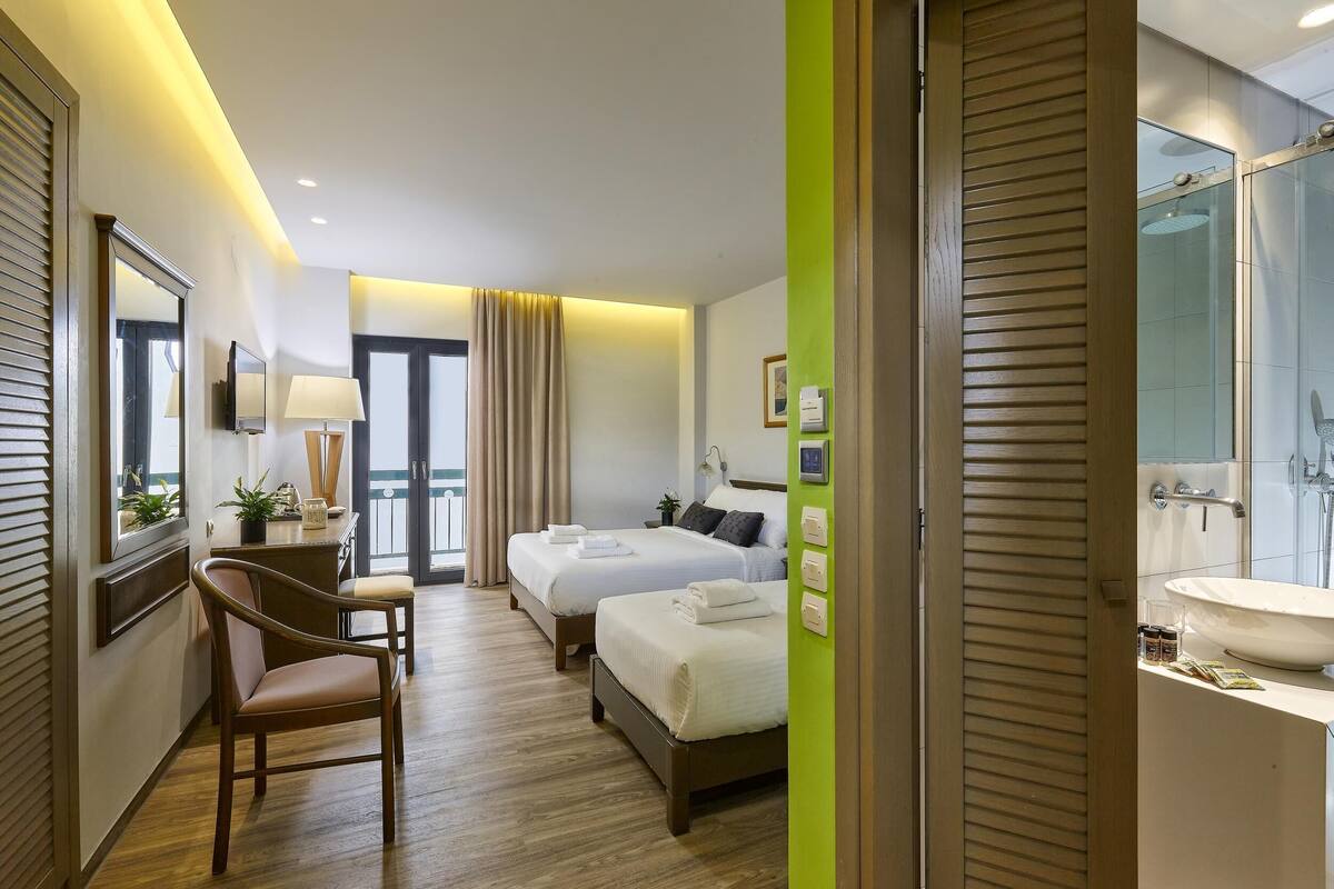 Habitación de hotel con dos camas, un escritorio, una silla, baño con lavamanos y ducha, y vista al exterior.