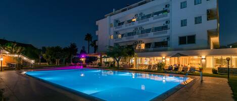 A swimming pool with a clear blue water surface, surrounded by a paved deck. There are multiple balconies on the adjacent building, and the area is illuminated by ambient lighting.