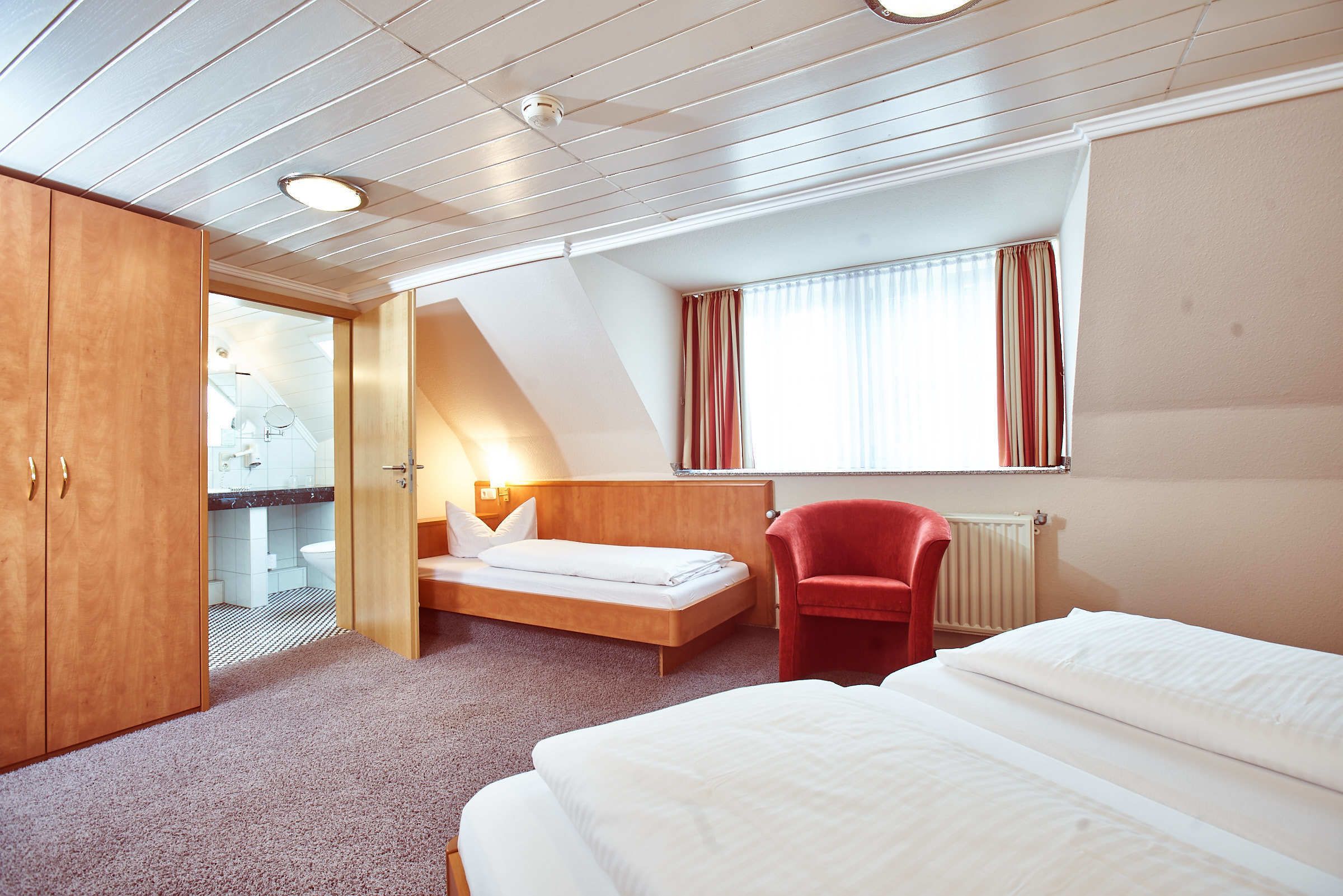 A hotel room with a wooden wardrobe, a bed, a red chair, a small desk, and a window with curtains.