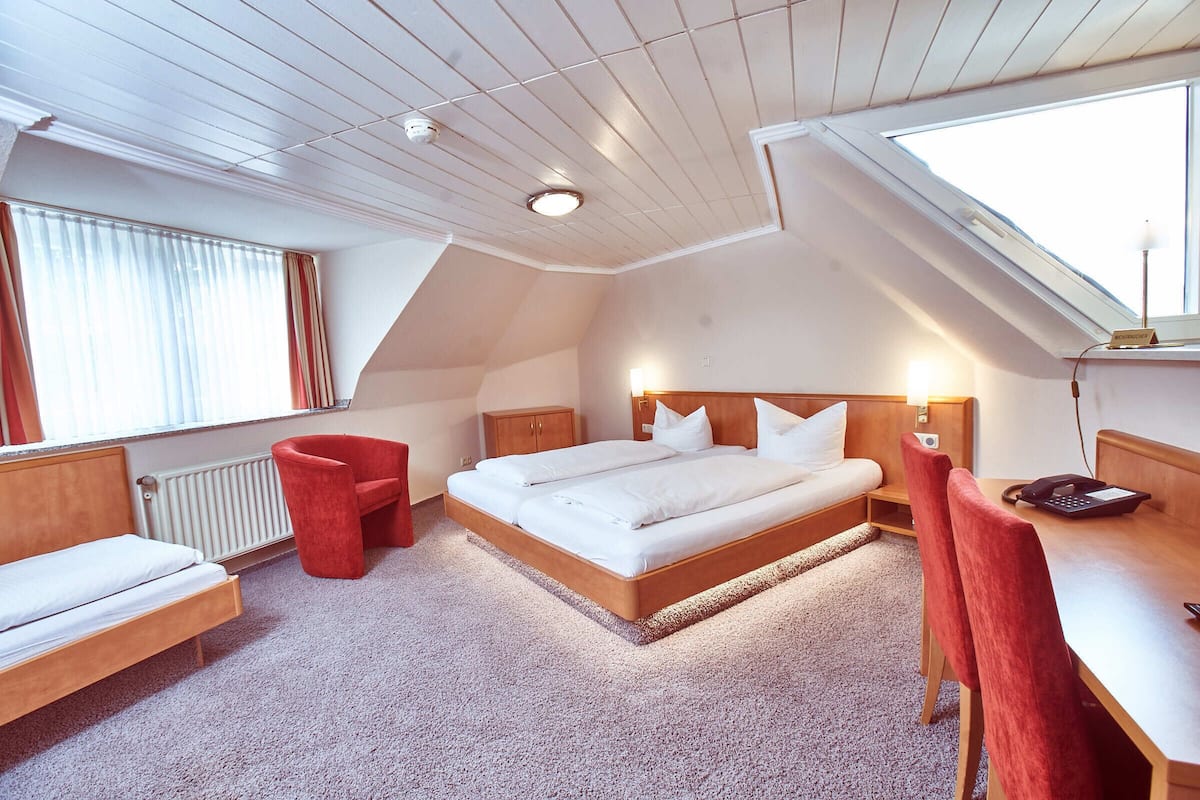 Habitación de hotel con una cama grande, un escritorio, una silla, un sillón rojo, una ventana con cortinas y un tragaluz.