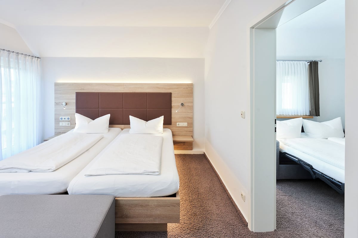 Habitación de hotel con dos camas, cabecera de madera, mesita de noche pequeña y vista al pasillo.