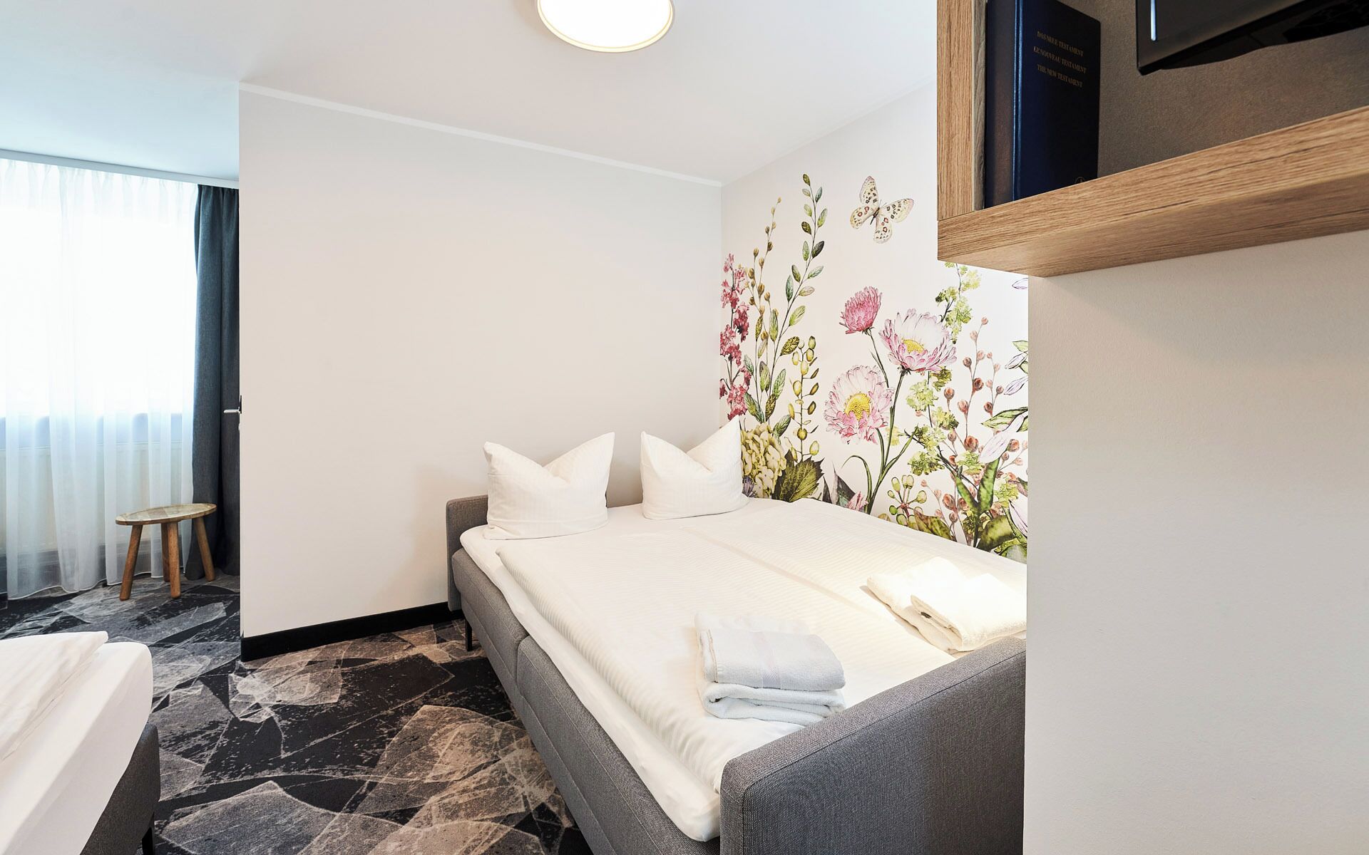 A hotel room with a bed, a small stool, a wall-mounted TV, and a floral wallpaper.