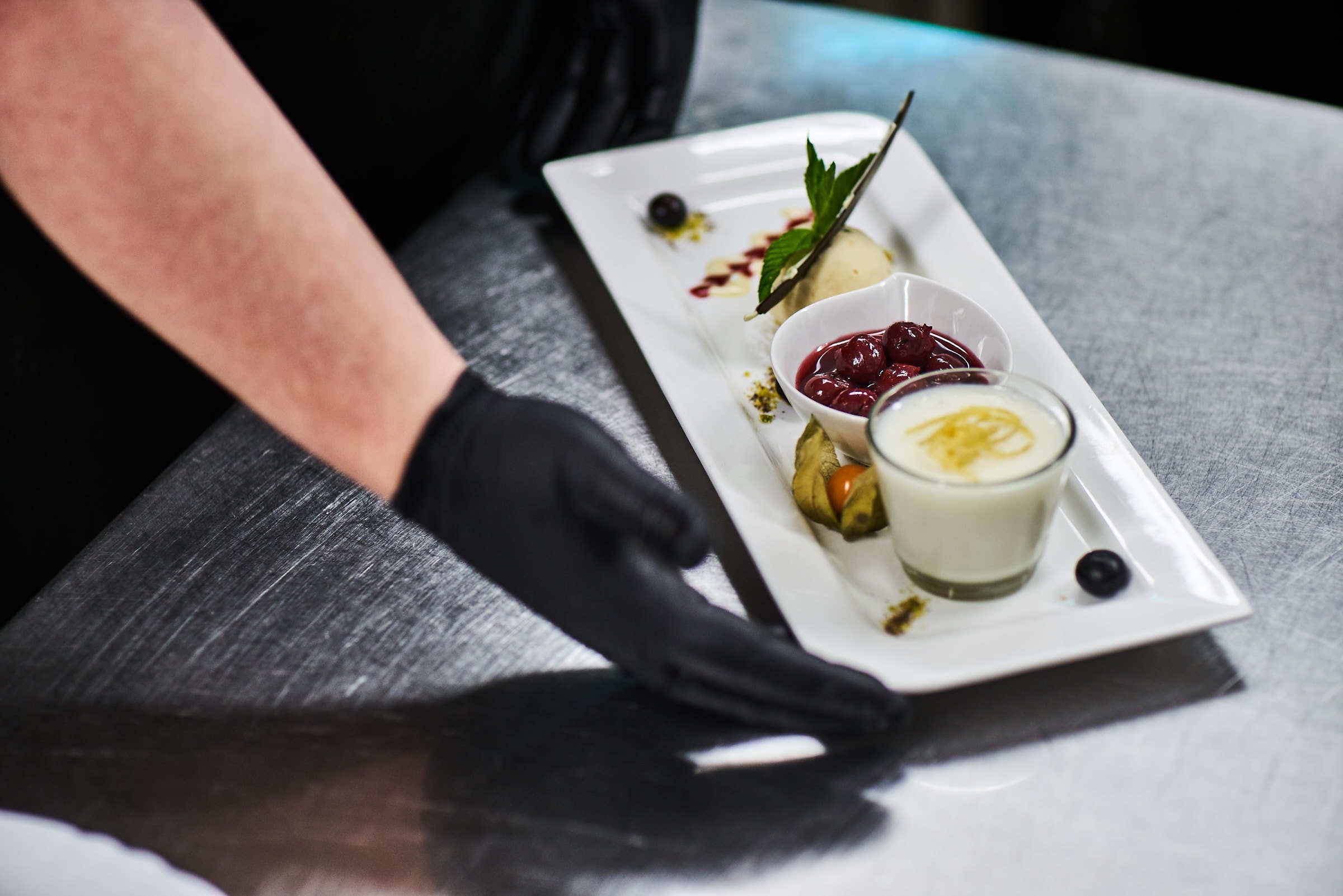 A white rectangular plate with various food items, including a dessert with a mint leaf garnish, a bowl of cherries, and a small glass of a creamy beverage.