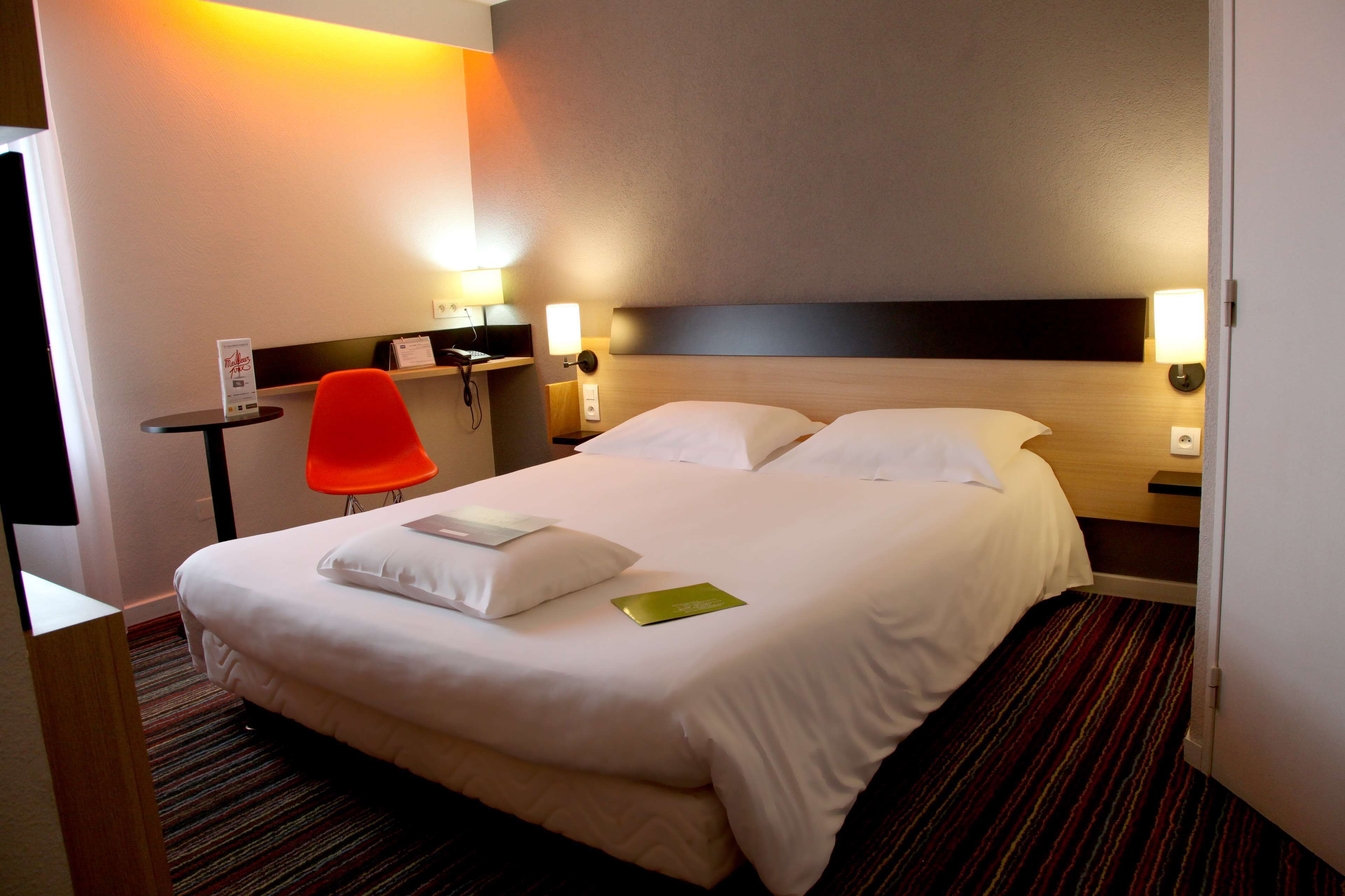 A hotel room with a large bed, two bedside tables with lamps, a small desk with a chair, and a red chair.