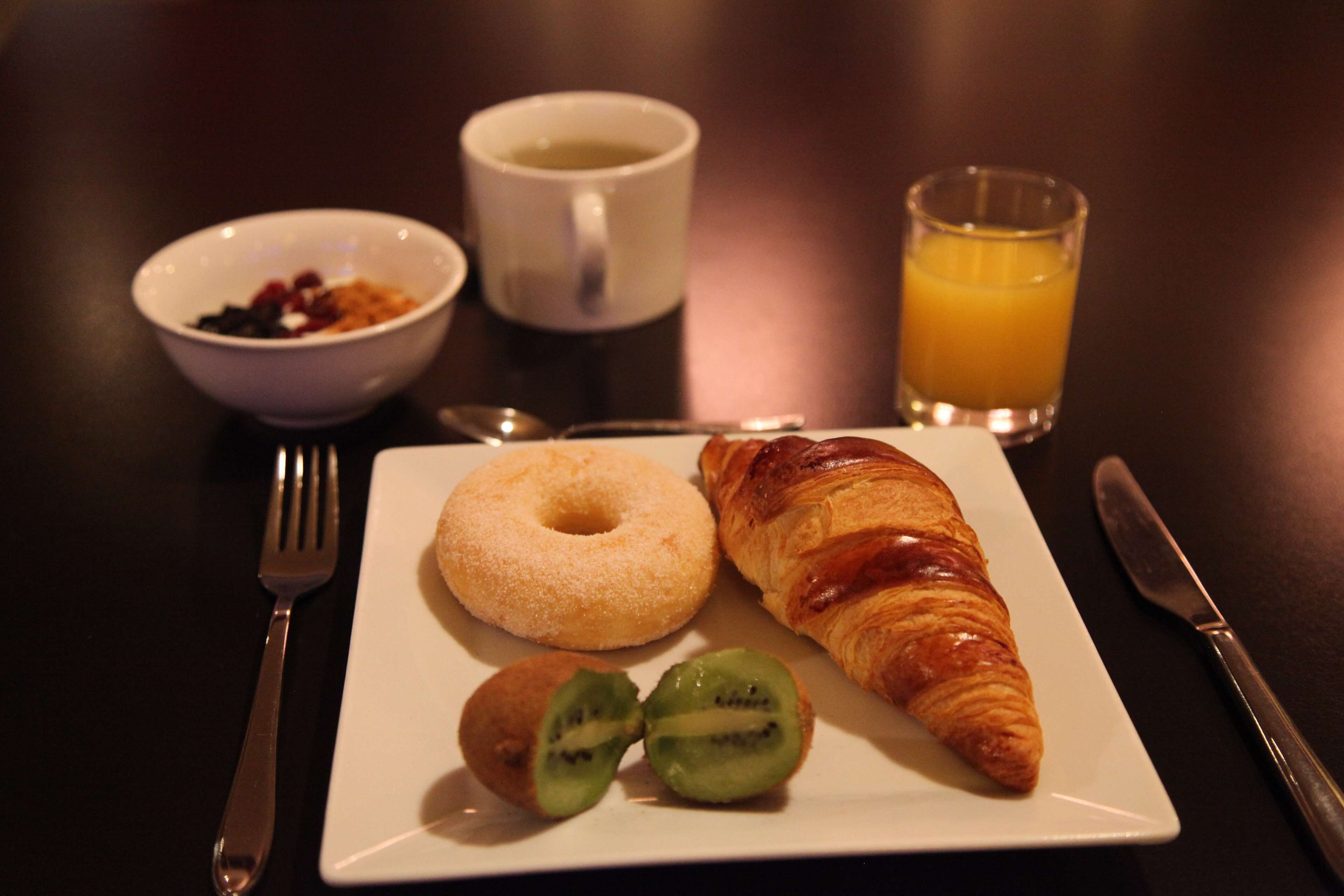 Prato de café da manhã com um croissant, um donut e fatias de kiwi, acompanhado de uma xícara de café e um copo de suco de laranja.
