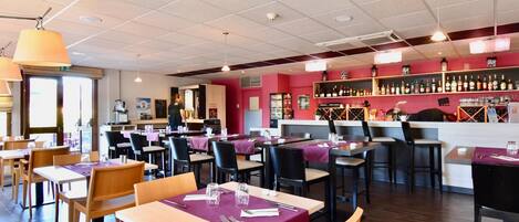 A restaurant interior with multiple dining tables, chairs, a bar area with shelves of bottles, and a ceiling with recessed lighting.