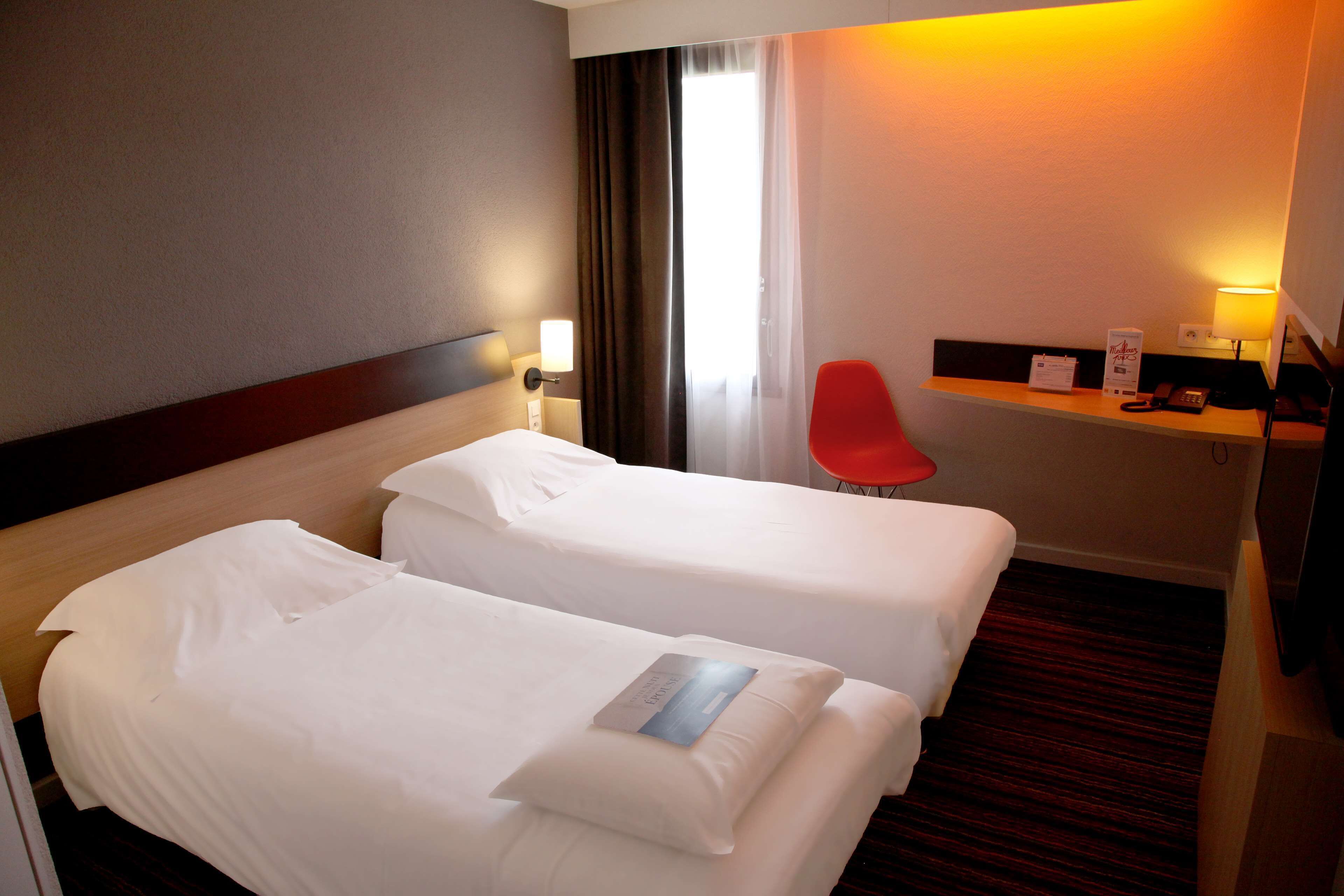 A hotel room with two beds, a desk with a lamp, a red chair, and a window with curtains.