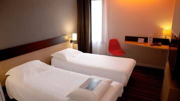 A hotel room with two beds, a desk with a lamp, a red chair, and a window with curtains.