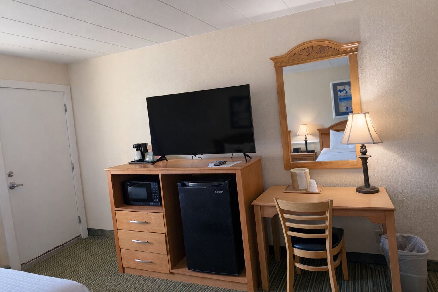 Una habitación de hotel con escritorio, televisión, microondas, refrigerador, silla, lámpara, basurero y cama.