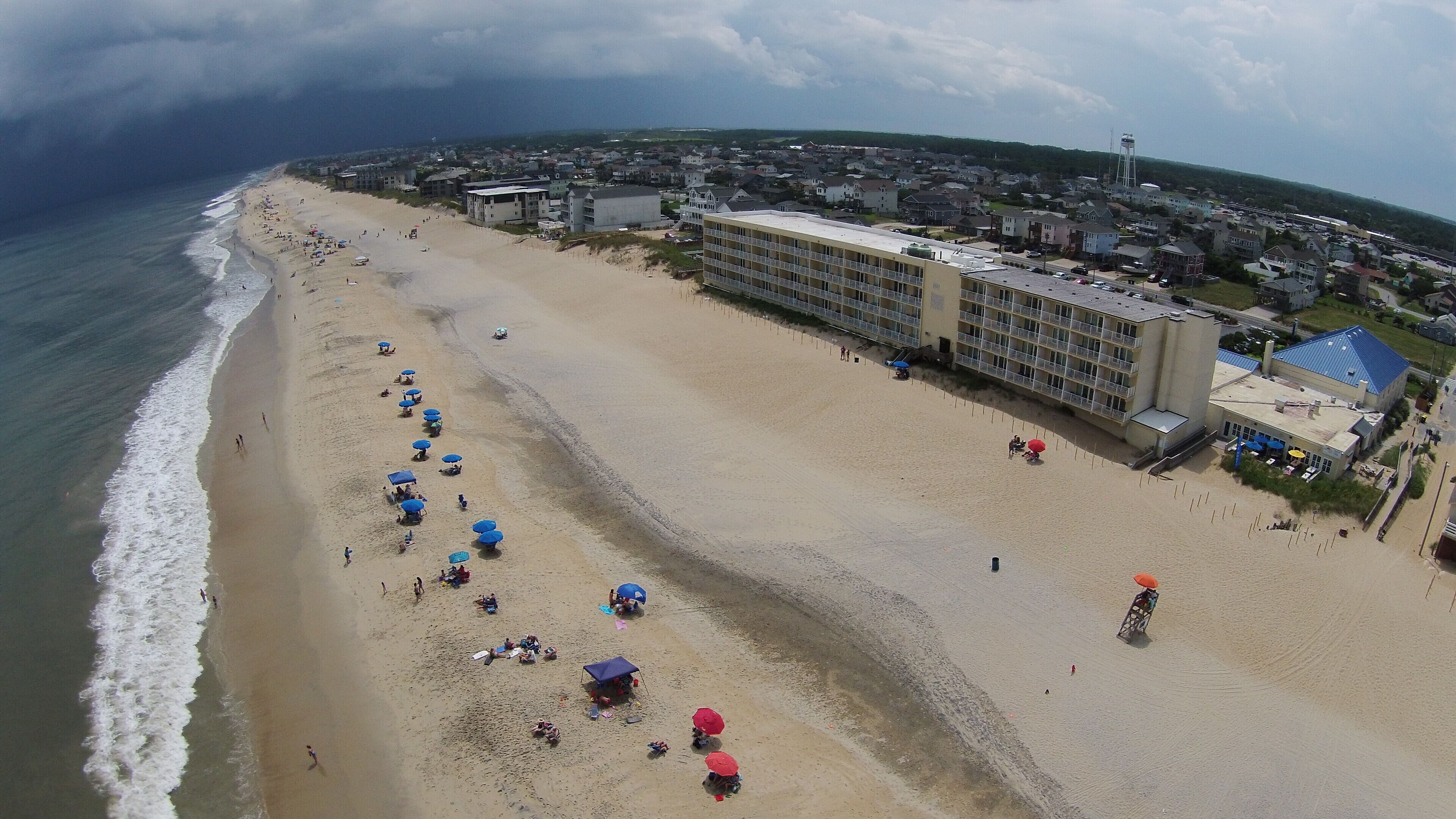 An aerial view of a beach with multiple beach umbrellas and a large hotel building with multiple floors and balconies.