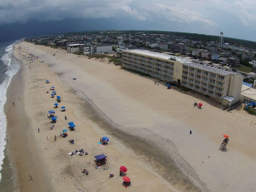 An aerial view of a beach with multiple beach umbrellas and a large hotel building with multiple floors and balconies.
