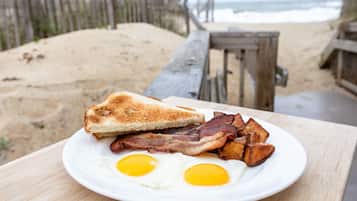 Une assiette de petit-déjeuner avec des toasts, du bacon, des œufs et des pommes de terre, posée sur une table en bois, avec une plage et une clôture en bois en arrière-plan.