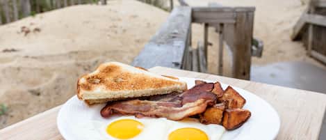 Un plato de desayuno con tostadas, tocino, huevos y papas sobre una mesa de madera, con una playa y una cerca de madera al fondo.