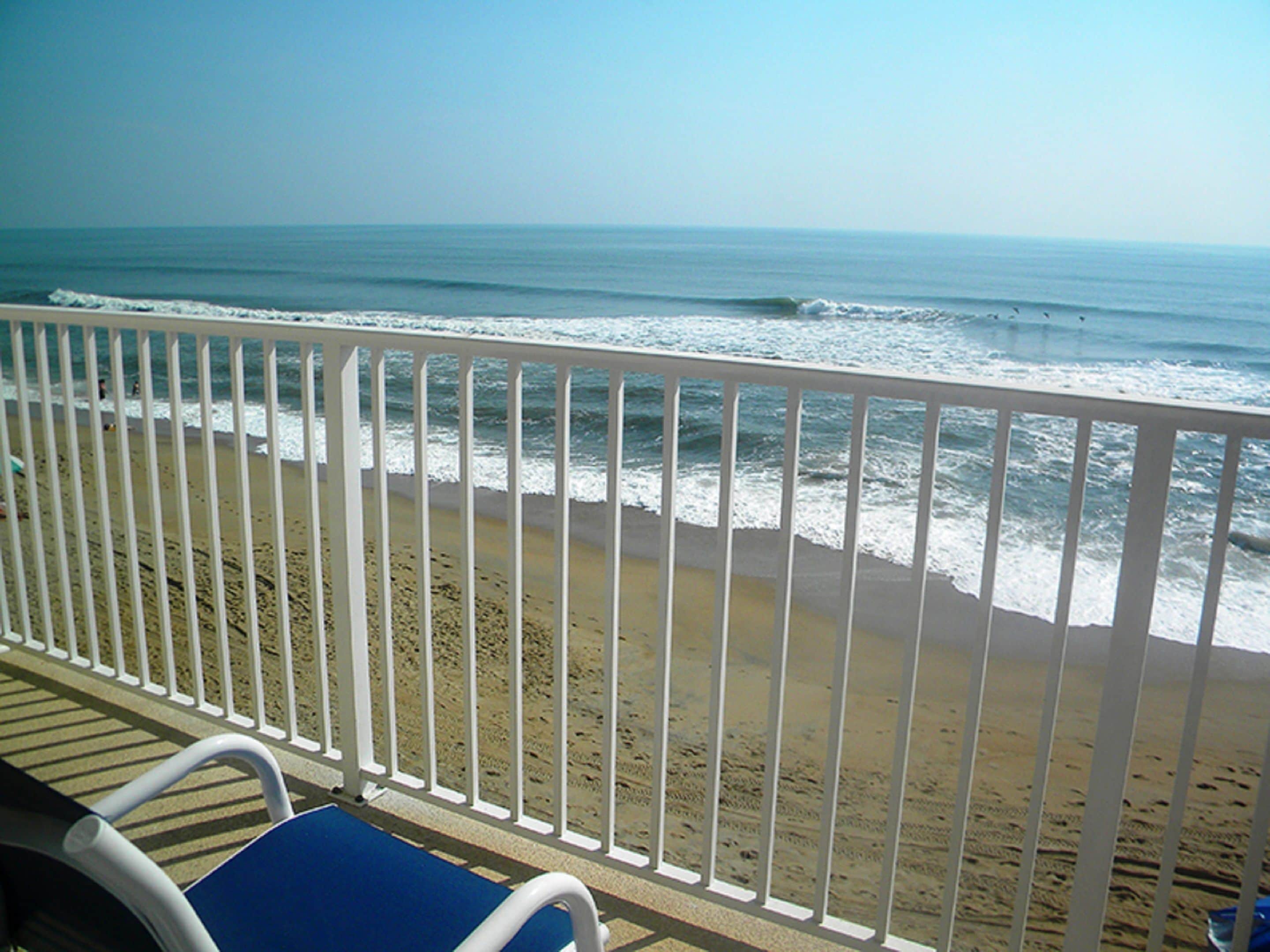 A beach view from a balcony with a white railing, a blue chair, and a sandy beach.