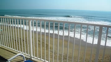 Vista a la playa desde un balcón con barandilla blanca, una silla azul y una playa arenosa.