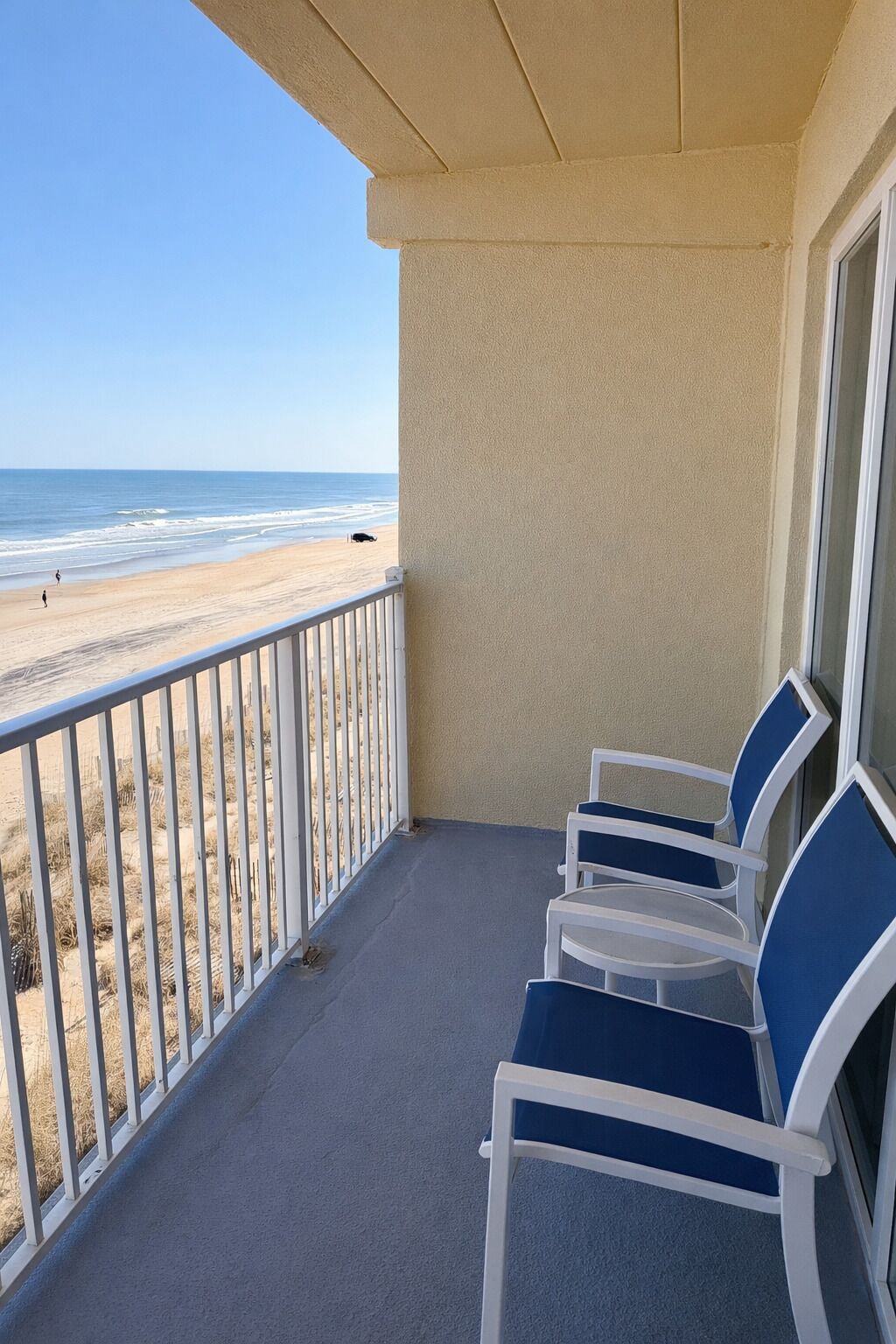 A balcony with a view of the beach, a railing, and two white chairs with blue cushions.