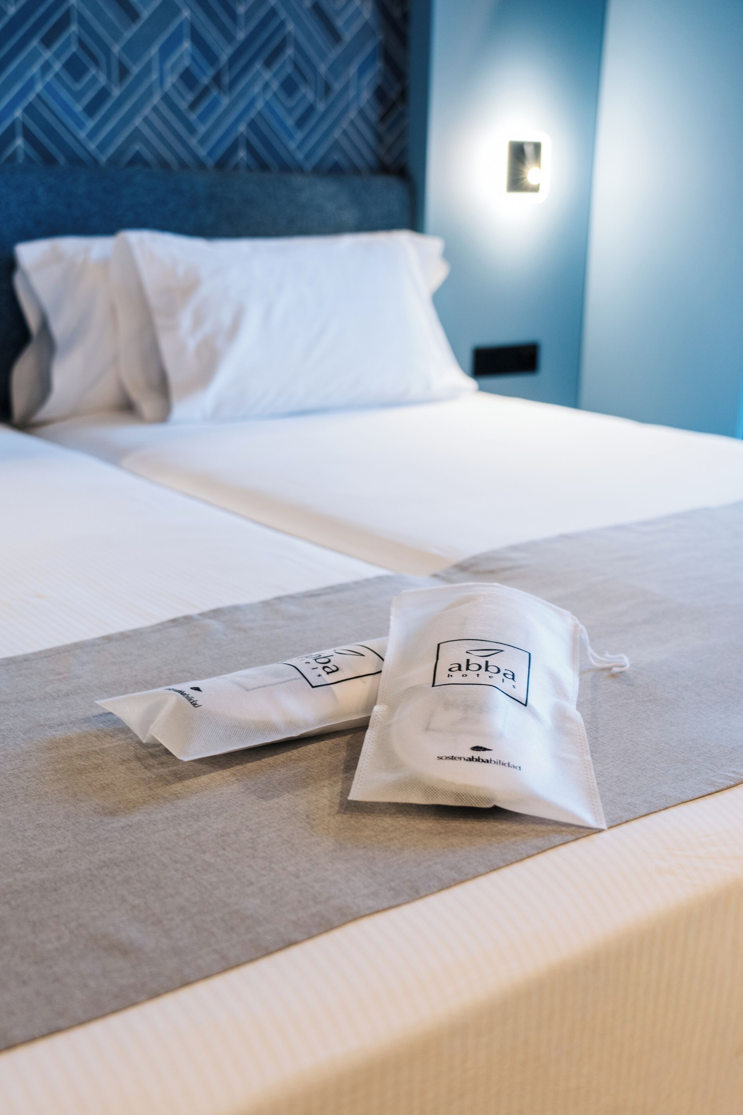 Una cama bien hecha con sábanas blancas y dos bolsas blancas con la etiqueta 'abba hotel' en un cabecero de madera.