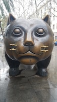 A large, metallic sculpture resembling a cat's face with a sad expression, positioned on a wet pavement in an urban setting.