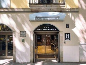 The entrance to HOTEL abba RAMBLAT with a clear glass facade, a sign indicating 24-hour service, and a welcome mat at the entrance.