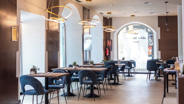 A modern restaurant with wooden tables and blue chairs, large windows, and decorative lighting. - Abba Rambla Barcelona (Barcelona)
